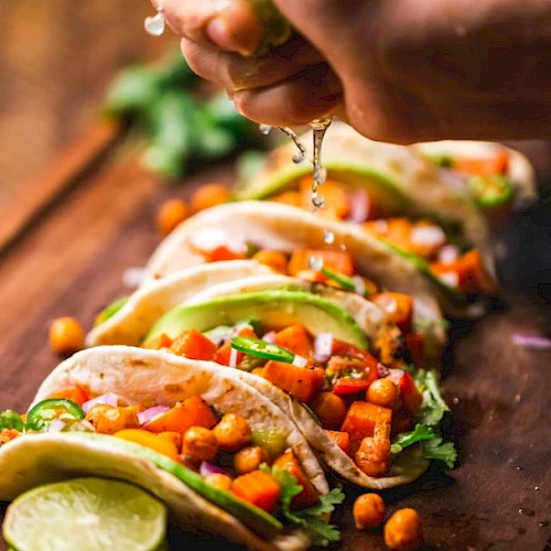 The image shows a person squeezing lime juice over a row of tacos filled with chickpeas, vegetables, and garnished with cilantro and avocado slices.