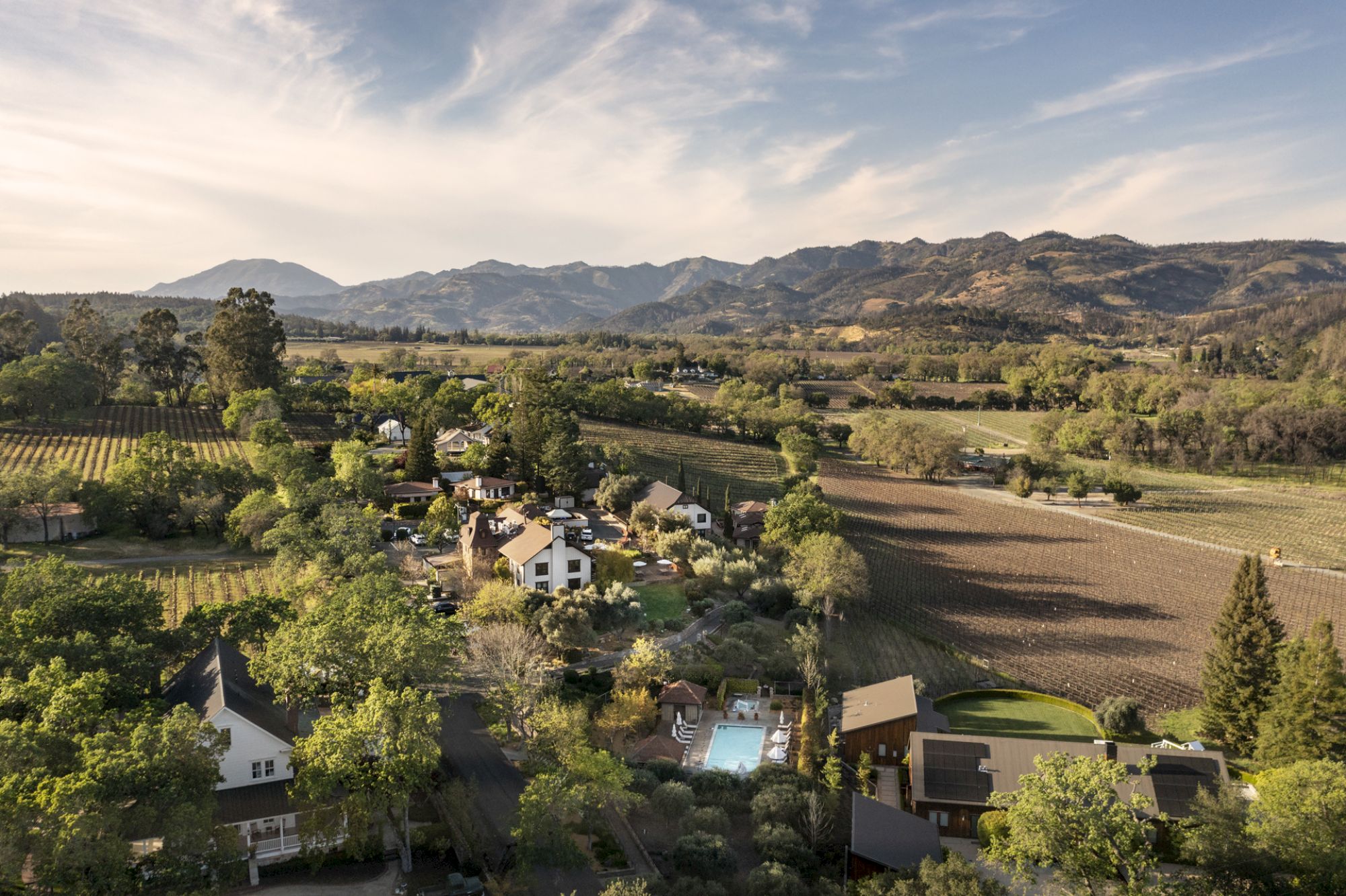 An aerial view of a picturesque village set amongst vineyards and lush greenery, surrounded by rolling hills and distant mountains.