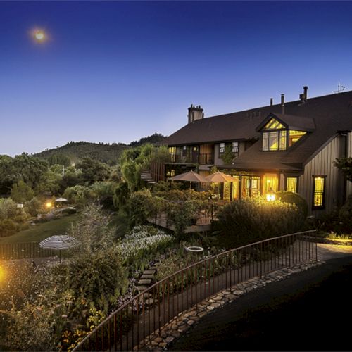 The image shows a large, illuminated house at dusk, surrounded by lush greenery and well-lit pathways, with a bright moon in the evening sky.