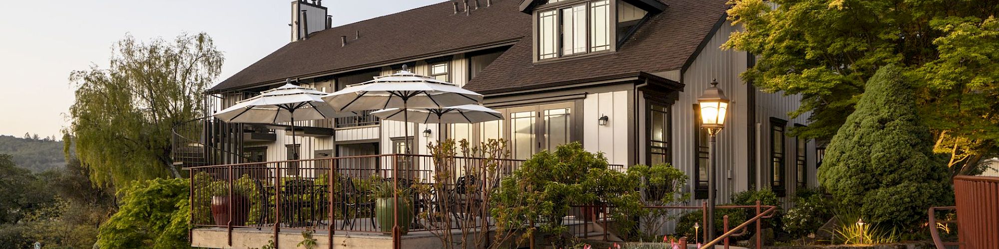 A beautiful house with a large deck featuring umbrellas and outdoor seating, surrounded by lush greenery and flowers, under a clear sky.