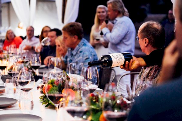 A group of people dining at a long table with wine glasses and flowers, while a man pours wine and another speaks into a microphone.