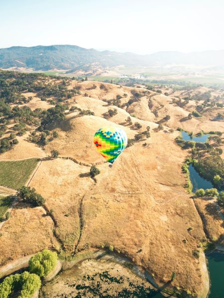 A colorful hot air balloon floats over a vast, hilly landscape with scattered trees, patches of green, and distant mountains in the background.