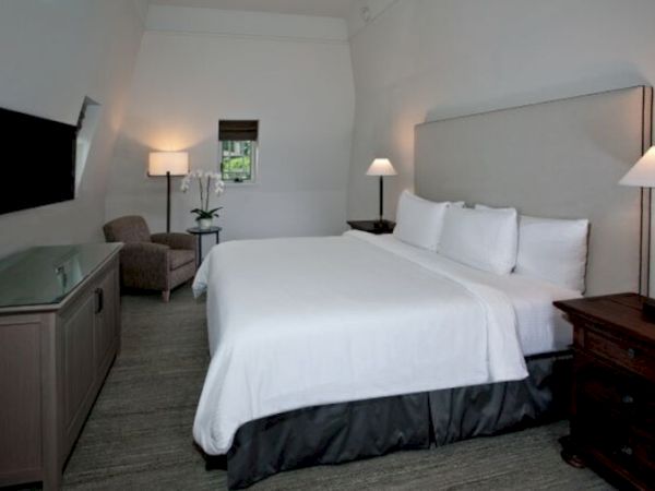 This image shows a modern hotel room with a large bed, two bedside tables with lamps, a flat-screen TV, an armchair, and a small window.