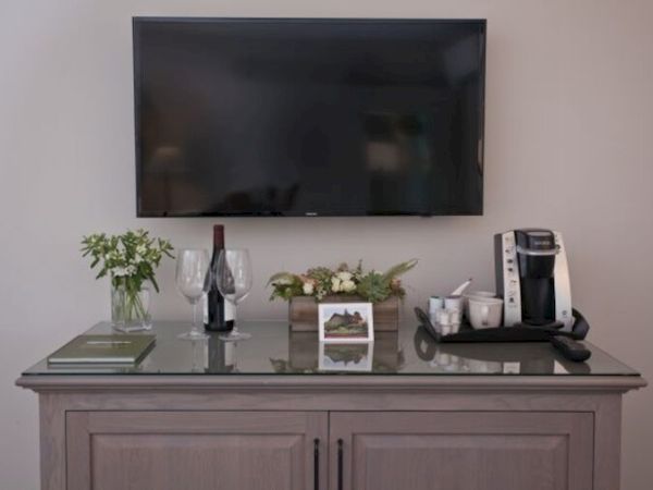 The image shows a wall-mounted TV above a cabinet with a plant, wine bottle, glasses, a coffee maker, and a small framed picture on it.