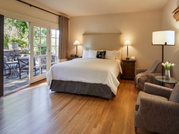 The image shows a cozy bedroom with a large bed, armchairs, a side table, and a doorway leading to an outdoor patio.