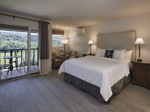 The image shows a well-lit hotel room with a large bed, two bedside tables, chairs, a balcony with a scenic view, and modern furnishings.