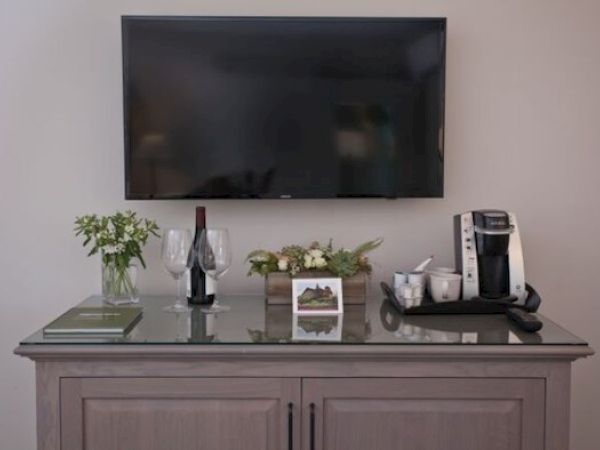 A wall-mounted TV above a cabinet with two wine glasses, a bottle, a coffee maker, a book, and some decorative plants on top.