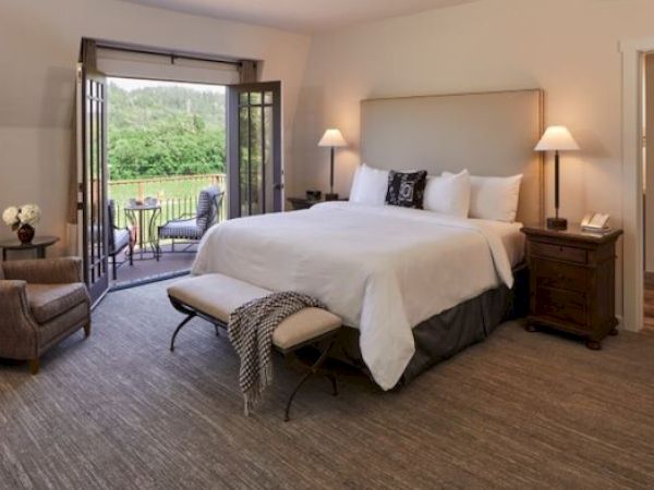 This image shows a cozy bedroom with a large bed, armchair, and open doors leading to an outdoor patio with seating overlooking a lush, green landscape.