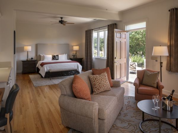 A cozy bedroom features a bed, sofa, armchair, desk, and table with wine glasses and a bottle, opening to a patio with greenery outside.