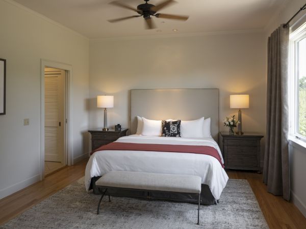 A neatly organized bedroom with a large bed flanked by two nightstands, each with a lamp, a bench at the foot of the bed, and a window to the right.