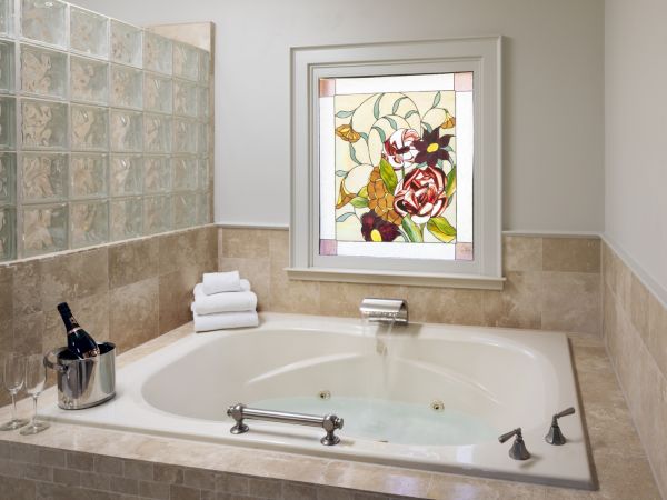 A luxurious bathroom with a large bathtub, towel stack, wine bottle with glasses, and a colorful stained glass window above the tub.