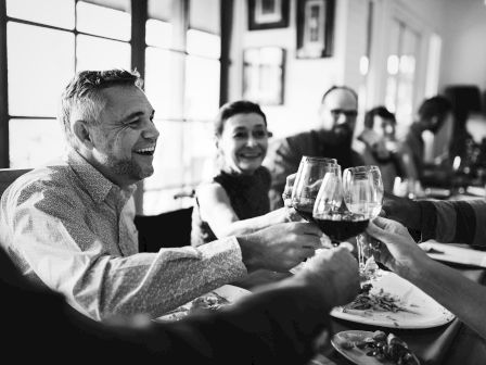 A group of people are sitting at a table, smiling, and clinking wine glasses in a celebratory toast.