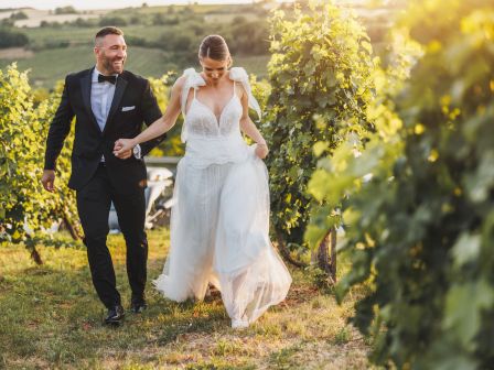 A couple dressed in wedding attire is walking hand in hand through a vineyard, smiling and enjoying the scenery at sunset.