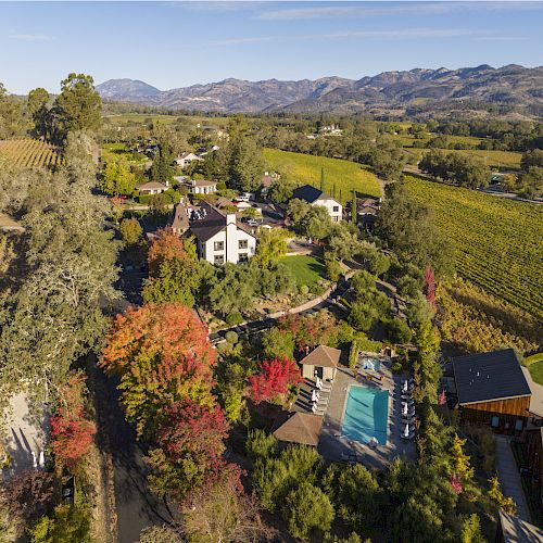 An aerial view of a picturesque area with vineyards, trees with colorful foliage, buildings, and a swimming pool surrounded by mountains.