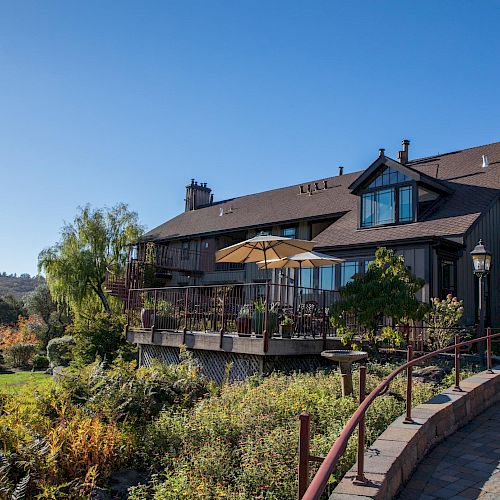 A beautiful, large house with a wooden exterior features a deck with outdoor seating and umbrellas, surrounded by lush greenery and scenic views.