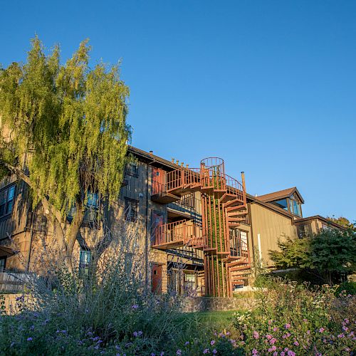 The image shows a multi-story building with a spiral staircase, surrounded by greenery and flowers, under a clear blue sky.