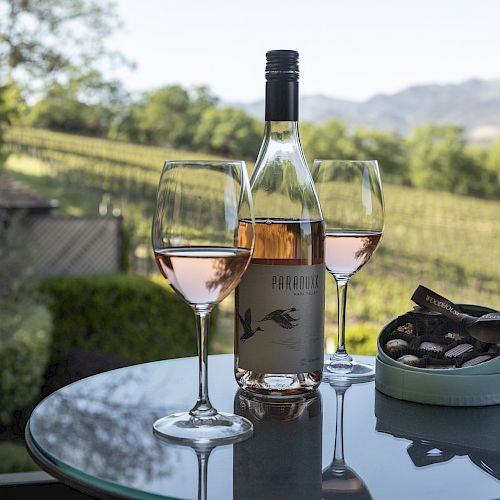 A bottle of rosé wine and two filled glasses sit on a glass table with a box of chocolates in an outdoor vineyard setting.