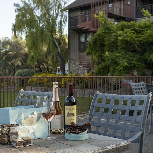 A table with wine bottles, glasses, a blue box, and snacks in an outdoor seating area with lush greenery and buildings in the background.