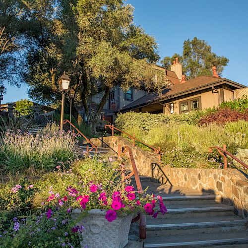 A stone path lined with vibrant flowers and greenery leading up to a charming, rustic house surrounded by tall trees and bathed in warm sunlight.