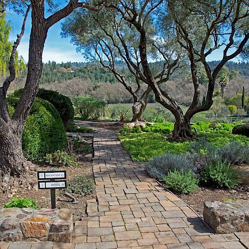 A stone pathway winds through a lush garden with trees, shrubs, and a sign indicating a visitor center and restrooms. Trees and hills are visible.