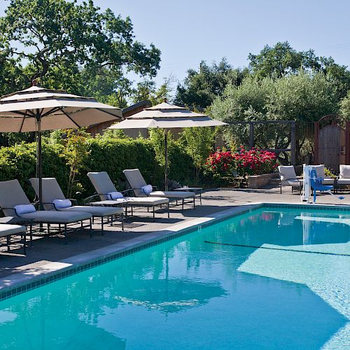 The image shows an outdoor swimming pool surrounded by lounge chairs with umbrellas, set in a garden area with trees and flowers.