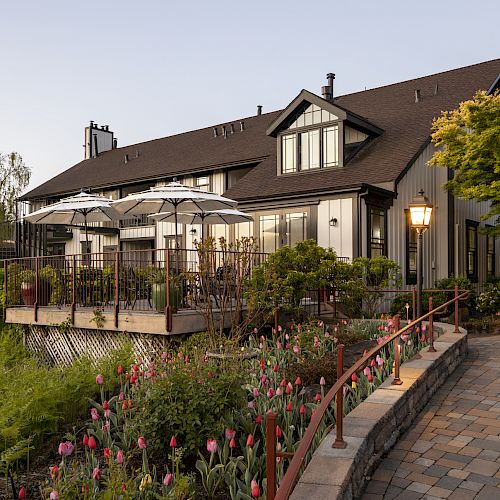 A two-story house with a patio equipped with tables and umbrellas, surrounded by a lush garden with blooming flowers and a brick pathway ending the sentence.