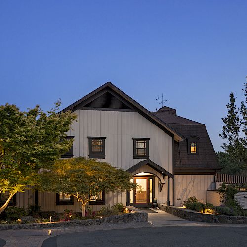 A charming two-story house set at dusk, surrounded by trees and well-maintained landscaping, with warm interior lights visible through windows.