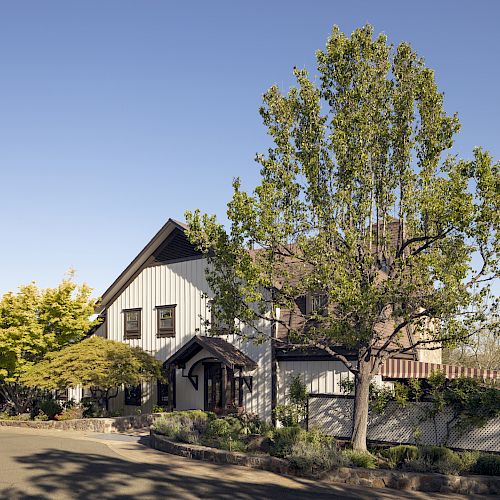 A two-story house with a gabled roof, surrounded by trees and plants, set against a clear blue sky, is shown in the image.