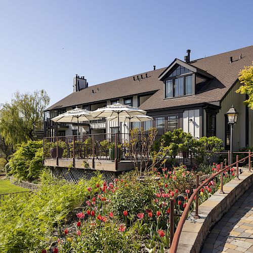 A beautiful two-story house with a stone path, red flowers, and outdoor umbrellas on a patio surrounded by lush greenery and blue sky.