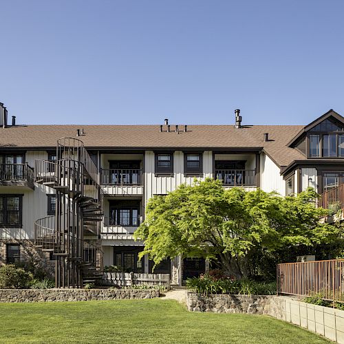 A multi-story house with a spiral staircase, surrounded by greenery, under a clear blue sky, featuring balconies and a well-maintained lawn.