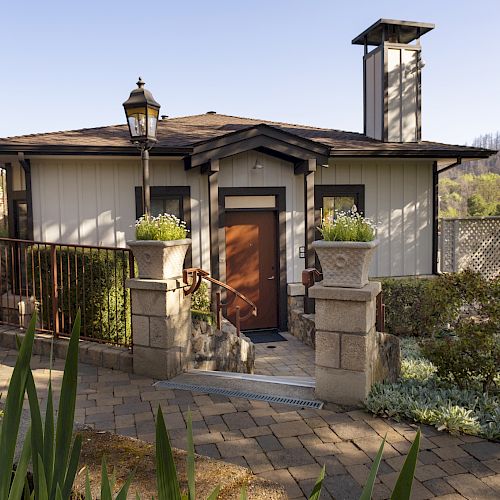 A quaint cottage with a wooden door, stone pillars, and lush greenery, framed by a cobblestone pathway and surrounded by trees in the background.