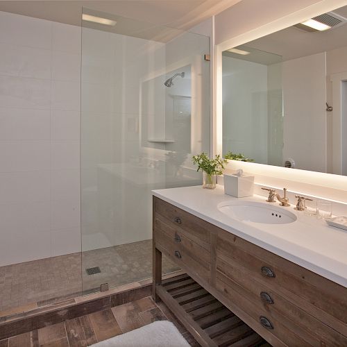 This image shows a modern bathroom with a glass-enclosed shower, a wooden vanity with a large mirror, sink, and various toiletries on the counter.