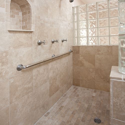 A walk-in shower with beige tiles, a built-in shelf, glass block wall, two showerheads, and a safety grab bar on the wall.