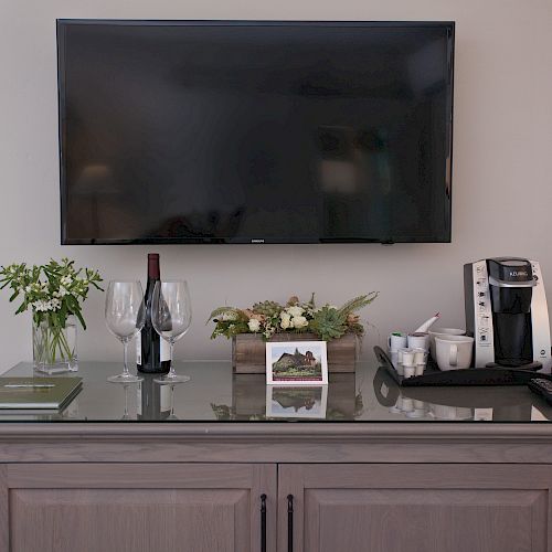 A flat-screen TV is mounted above a console with flowers, wine, glasses, a book, a floral arrangement, and a coffee maker with cups.