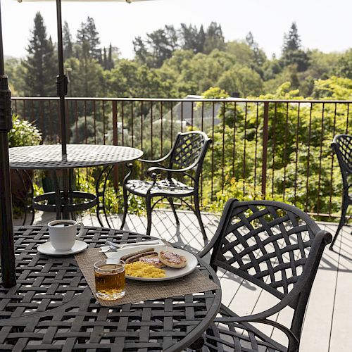 A patio with black wrought iron furniture, featuring a table set with a meal including eggs, toast, and a drink, overlooking a lush scenic view.