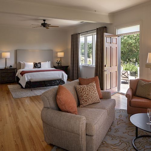 A cozy bedroom with a bed, bedside tables, a sofa, chairs, a desk, and an open door to a balcony. Wine and glasses are on a small table.