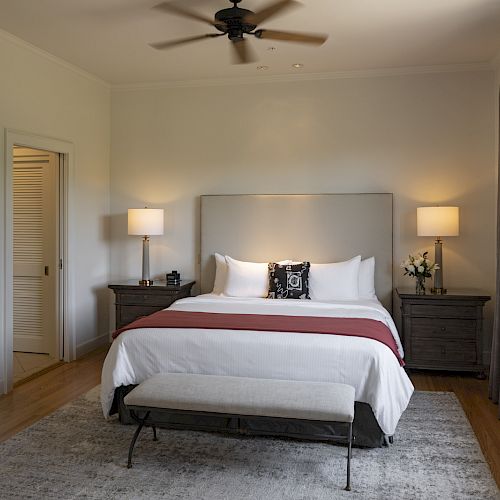 A modern bedroom with a king-size bed, two nightstands with lamps, a ceiling fan, a bench at the foot of the bed, and a large window with curtains.