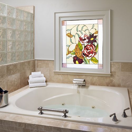 A luxurious bathtub with light-colored tiles, a wine bottle in an ice bucket, two glasses, and towels, adorned with a floral stained glass window.