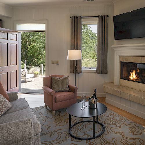 A cozy living room with a couch, armchair, coffee table with wine, fireplace, wall-mounted TV, window views, open door, and natural light ending the sentence.