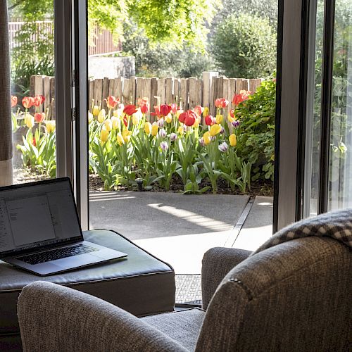 A cozy indoor corner with a chair, a plaid blanket, and a laptop on an ottoman. The open door reveals a garden with blooming tulips and greenery.