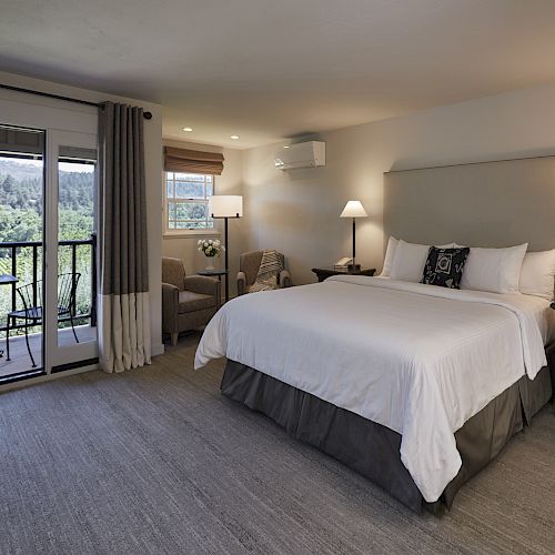 A spacious bedroom features a large bed, bedside tables with lamps, a sitting area, and an open door leading to a balcony with outdoor furniture.