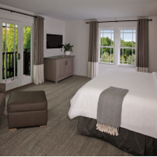 A cozy bedroom featuring a large bed, chair, ottoman, a TV on a cabinet, windows with a scenic view, and a door leading to a balcony.