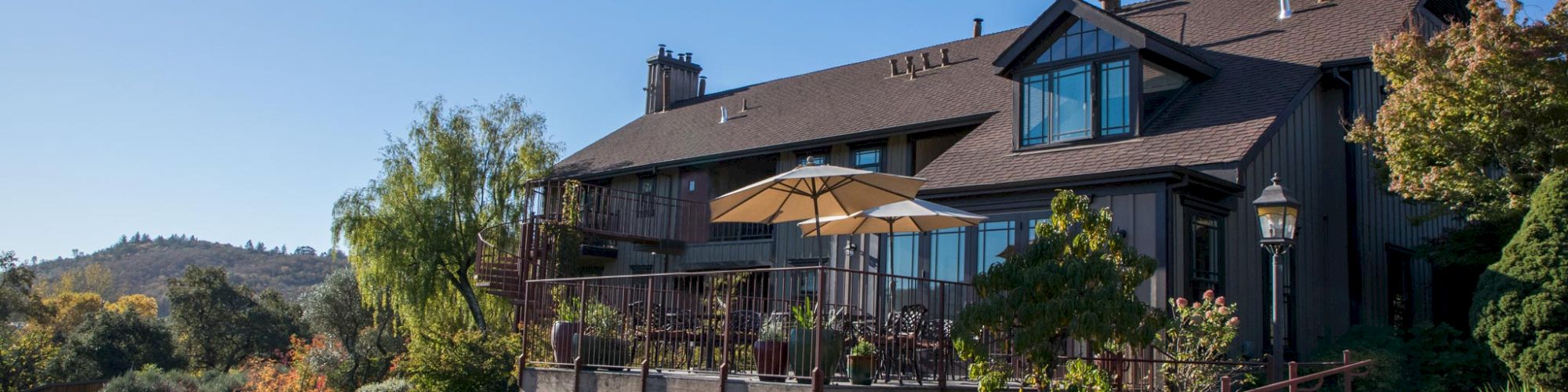 A large house with a wooden exterior sits amid lush greenery, featuring a patio with tables and umbrellas, under a clear blue sky.