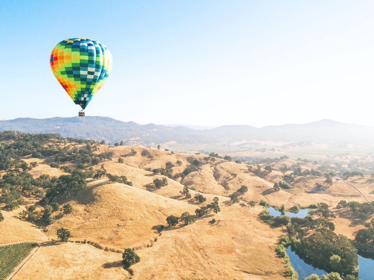 A colorful hot air balloon floats over a scenic landscape of rolling hills, sparse trees, and small lakes under a clear blue sky.